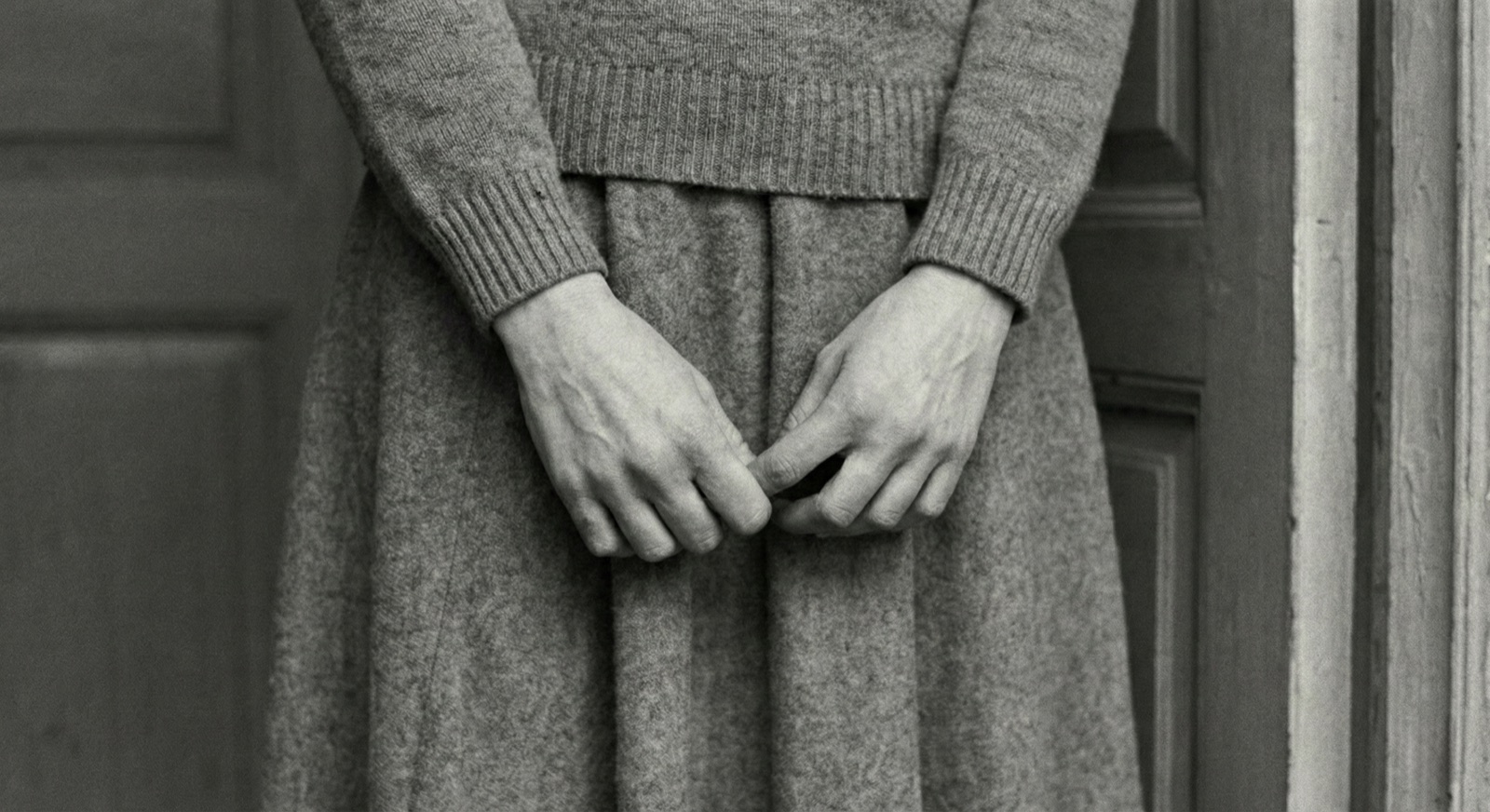 Hana's clasped hands over a grey wool skirt, in the doorway