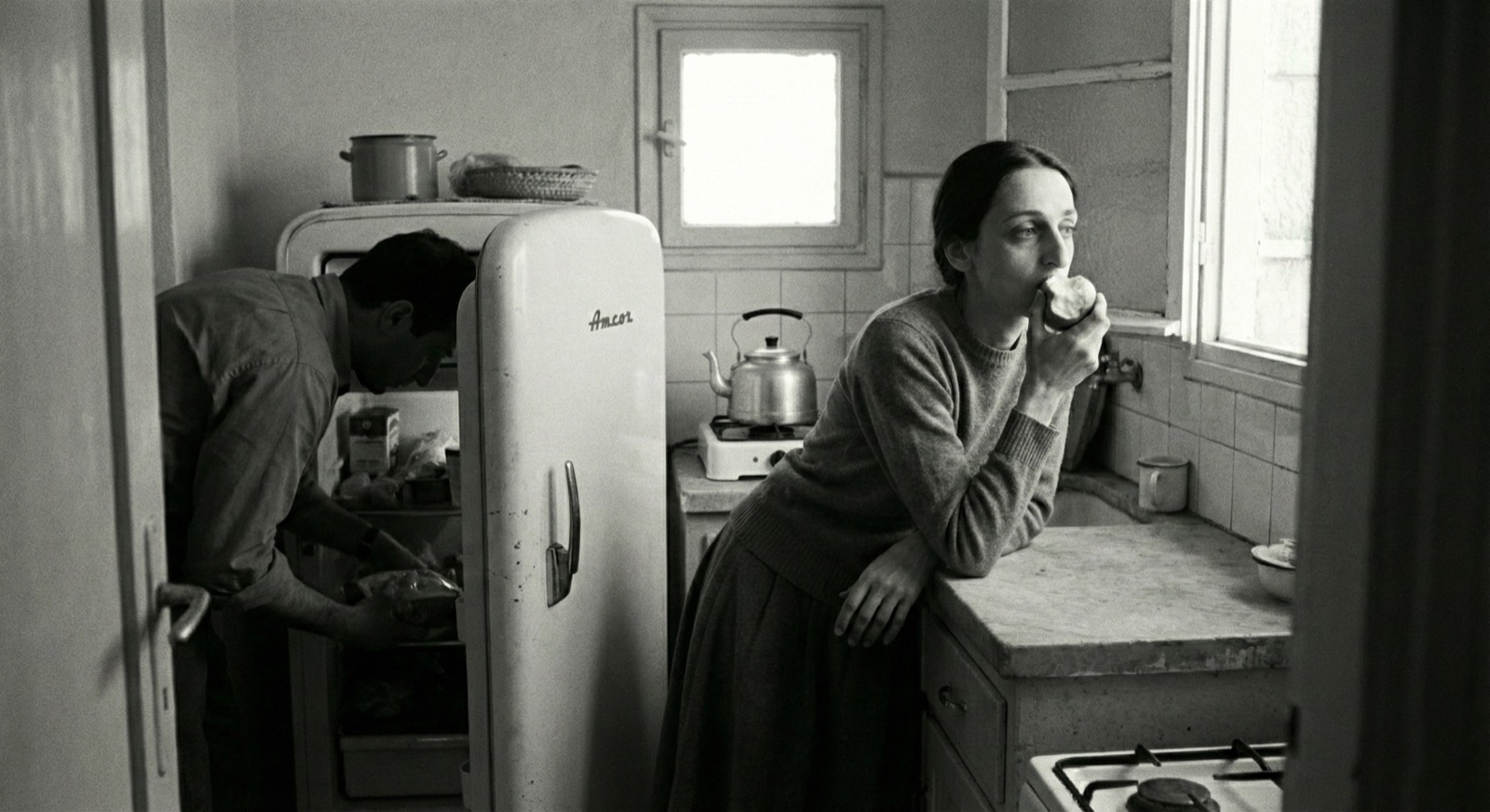 Hana eating an apple at the kitchen window while Michael bends into the refrigerator