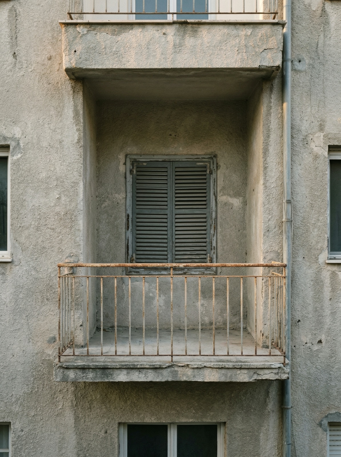 An empty concrete balcony with a rusted railing and closed grey shutters
