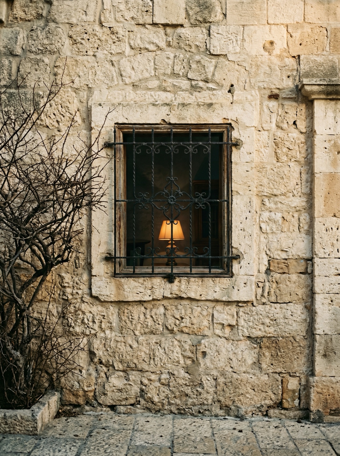 A single lamp glowing behind a barred window in a Jerusalem stone facade